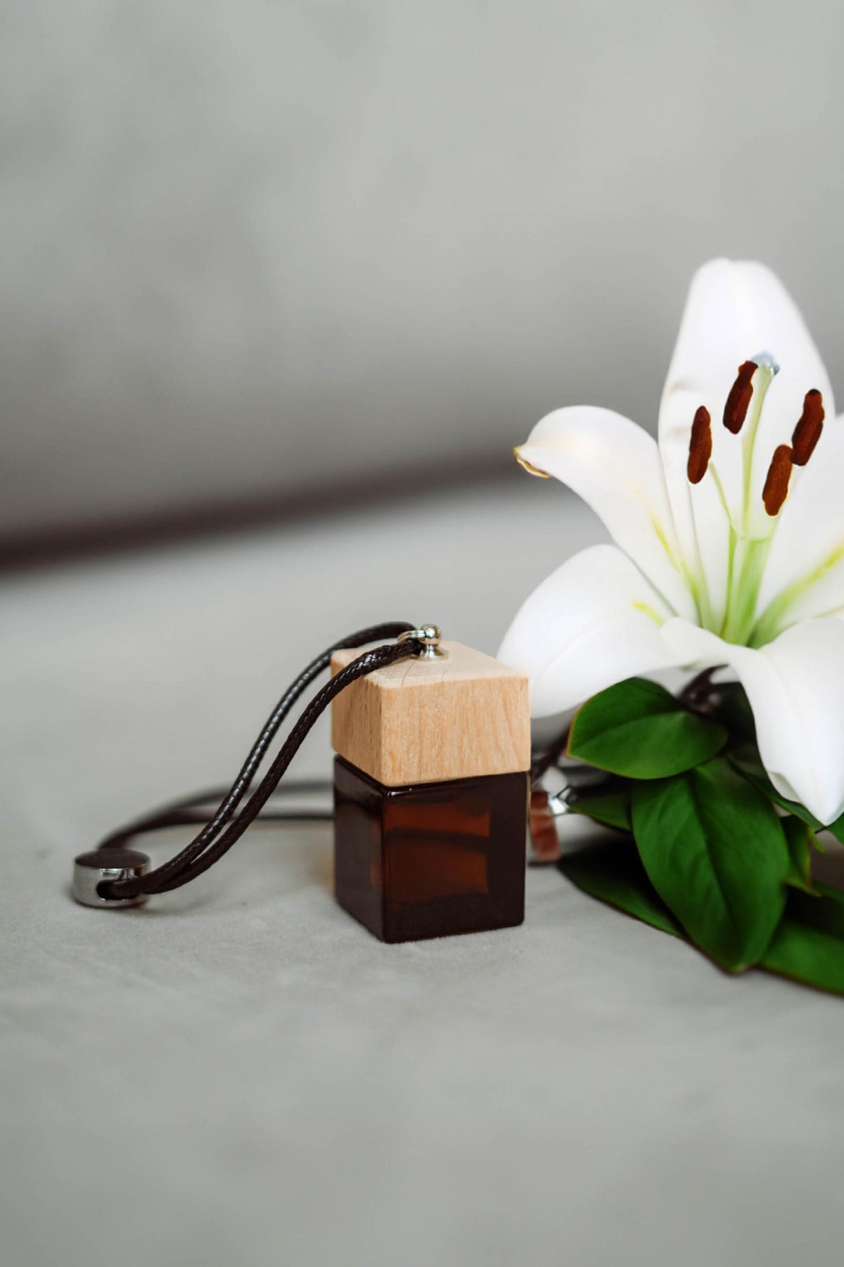 Άρωμα αυτοκινήτου Bamboo BY VELCHEVA 10ml σε γυάλινο μπουκάλι amber με ξύλινο καπάκι και δερμάτινο κορδόνι, δίπλα σε λευκό κρίνο
