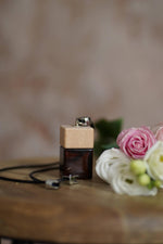 Άρωμα αυτοκινήτου Fleur BY VELCHEVA 10ml σε γυάλινο μπουκάλι amber με ξύλινο καπάκι και μαύρο κορδόνι, δίπλα σε ροζ και λευκά τριαντάφυλλα