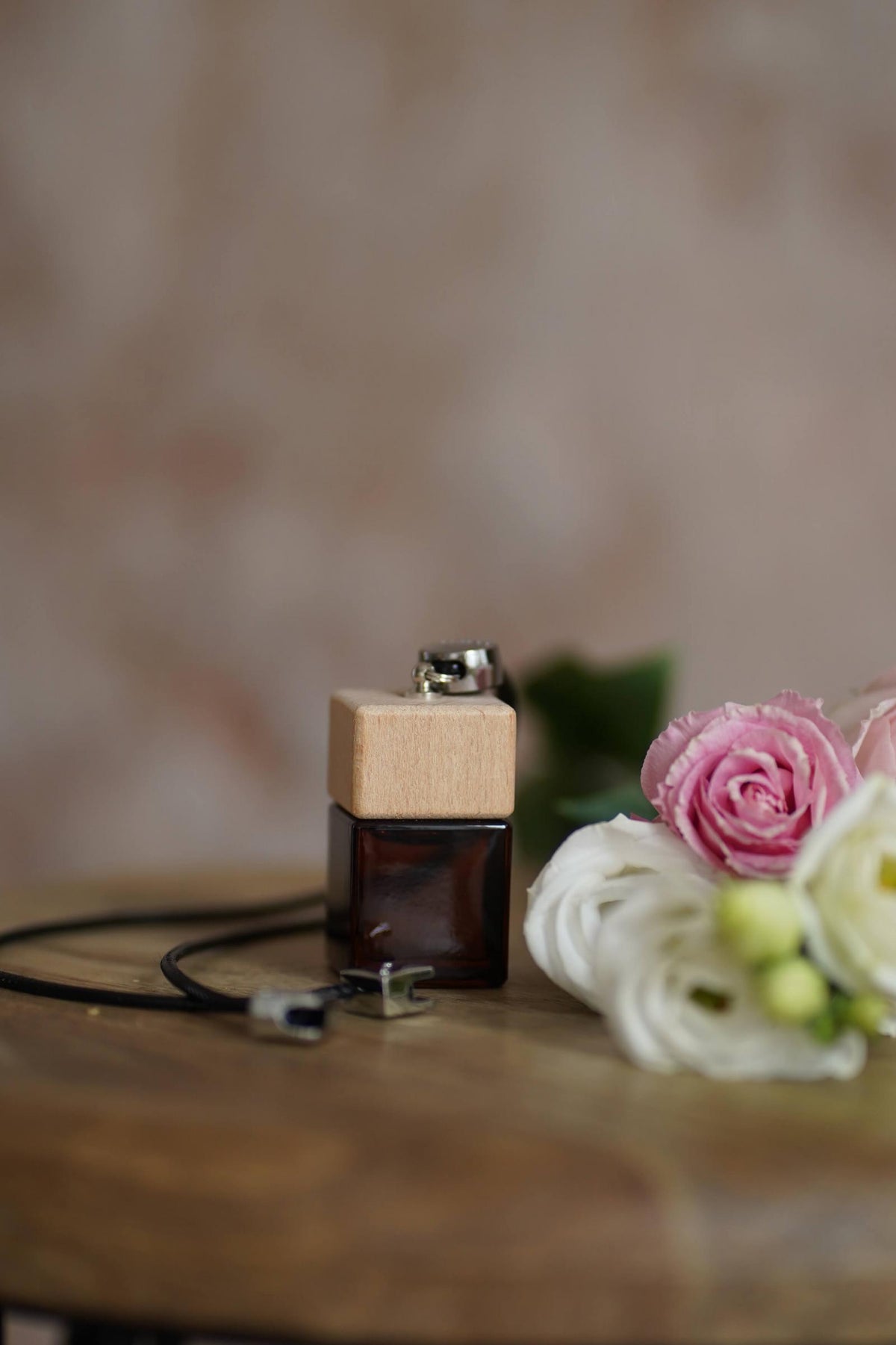 Άρωμα αυτοκινήτου Fleur BY VELCHEVA 10ml σε γυάλινο μπουκάλι amber με ξύλινο καπάκι και μαύρο κορδόνι, δίπλα σε ροζ και λευκά τριαντάφυλλα