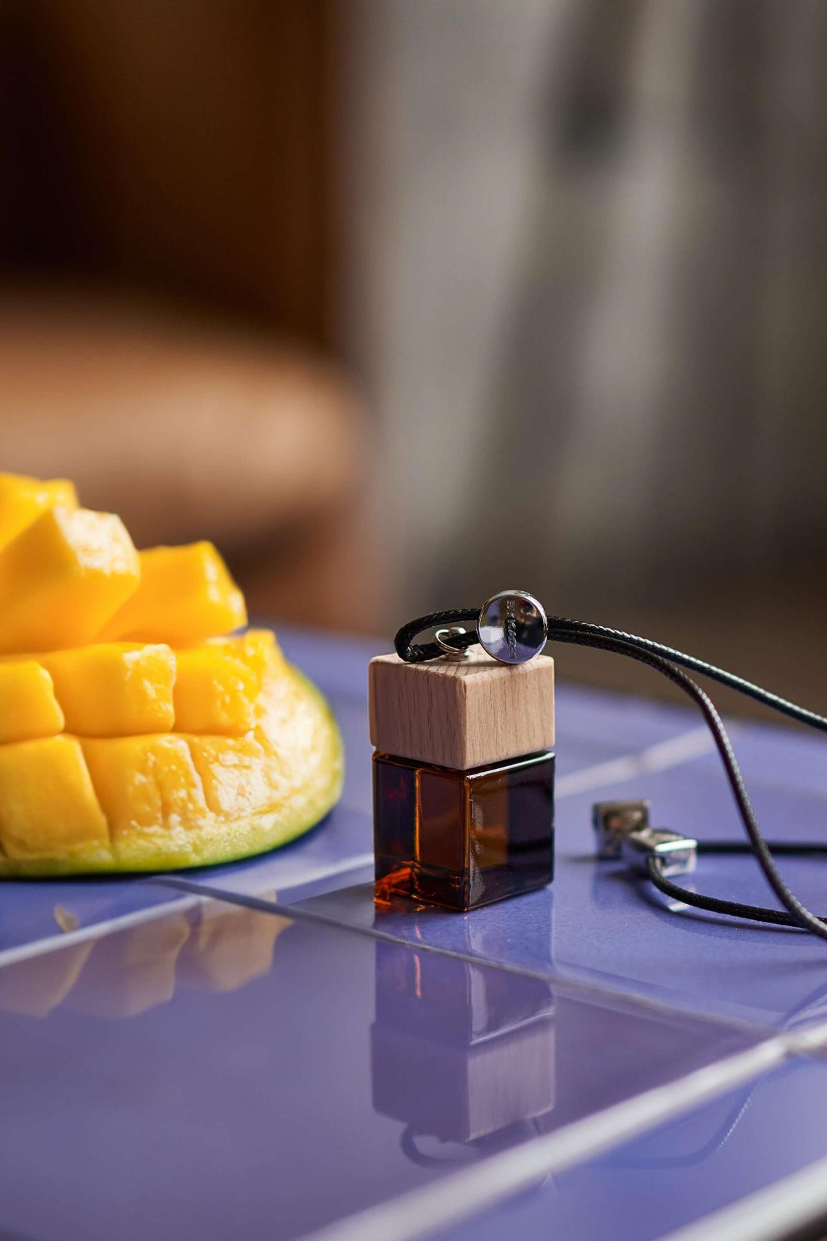 Άρωμα αυτοκινήτου Green Mango BY VELCHEVA 10ml σε γυάλινο μπουκάλι amber με ξύλινο καπάκι και μαύρο κορδόνι, δίπλα σε κομμένο φρέσκο μάνγκο