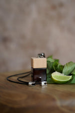 Άρωμα αυτοκινήτου Lime Basil BY VELCHEVA 10ml σε γυάλινο μπουκάλι amber με ξύλινο καπάκι και μαύρο κορδόνι, δίπλα σε λάιμ και φύλλα βασιλικού