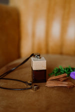 Άρωμα αυτοκινήτου Santal BY VELCHEVA 10ml σε γυάλινο μπουκάλι amber με ξύλινο καπάκι και μαύρο κορδόνι, δίπλα σε ξύσματα σανδαλόξυλου