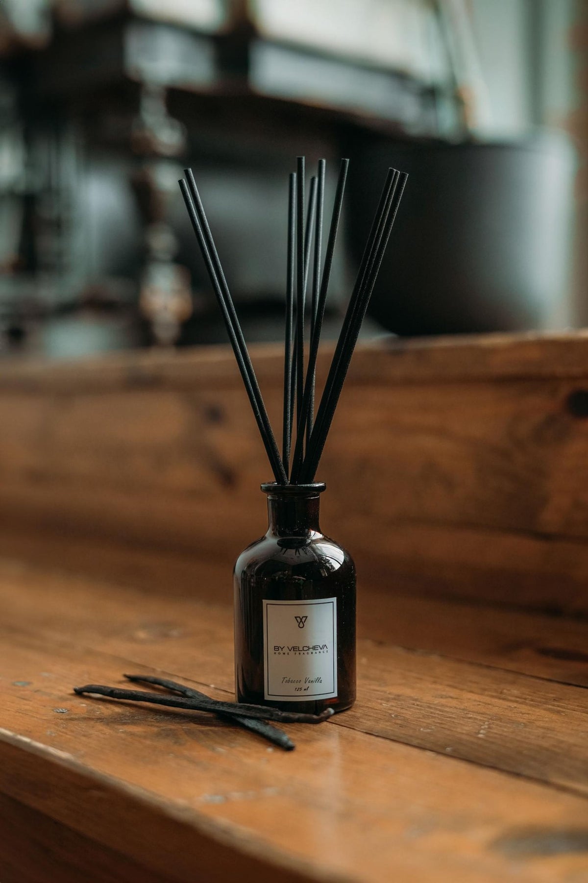 Αρωματικό χώρου με στικ Tobacco Vanilla BY VELCHEVA σε καφέ γυάλινο μπουκάλι πάνω σε ξύλινη επιφάνεια, με λοβούς βανίλιας 125 ml
