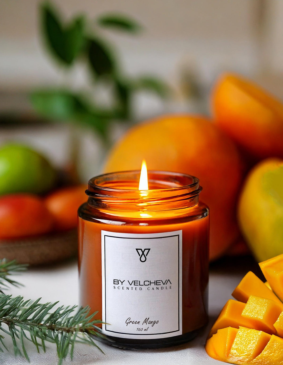 Αρωματικό κερί Green Mango BY VELCHEVA 160ml σε γυάλινο βάζο amber, αναμμένο, με ολόκληρα και κομμένα μάνγκο σε φωτεινό φόντο