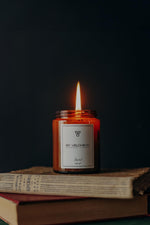 Αρωματικό κερί Santal BY VELCHEVA 160ml σε γυάλινο βάζο amber, αναμμένο, πάνω σε παλιά βιβλία σε σκούρο φόντο
