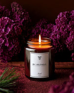 Αρωματικό κερί Santal BY VELCHEVA 160ml σε γυάλινο βάζο amber, αναμμένο, με μοβ πασχαλιές και κλαδί πεύκου
