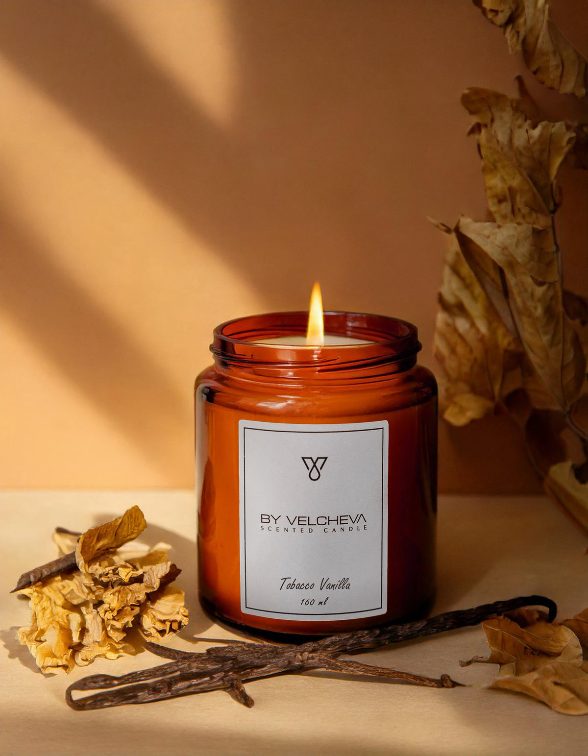 Αρωματικό κερί Tobacco Vanilla BY VELCHEVA 160ml σε γυάλινο βάζο amber, αναμμένο, με φύλλα καπνού και λοβούς βανίλιας σε ζεστό φόντο