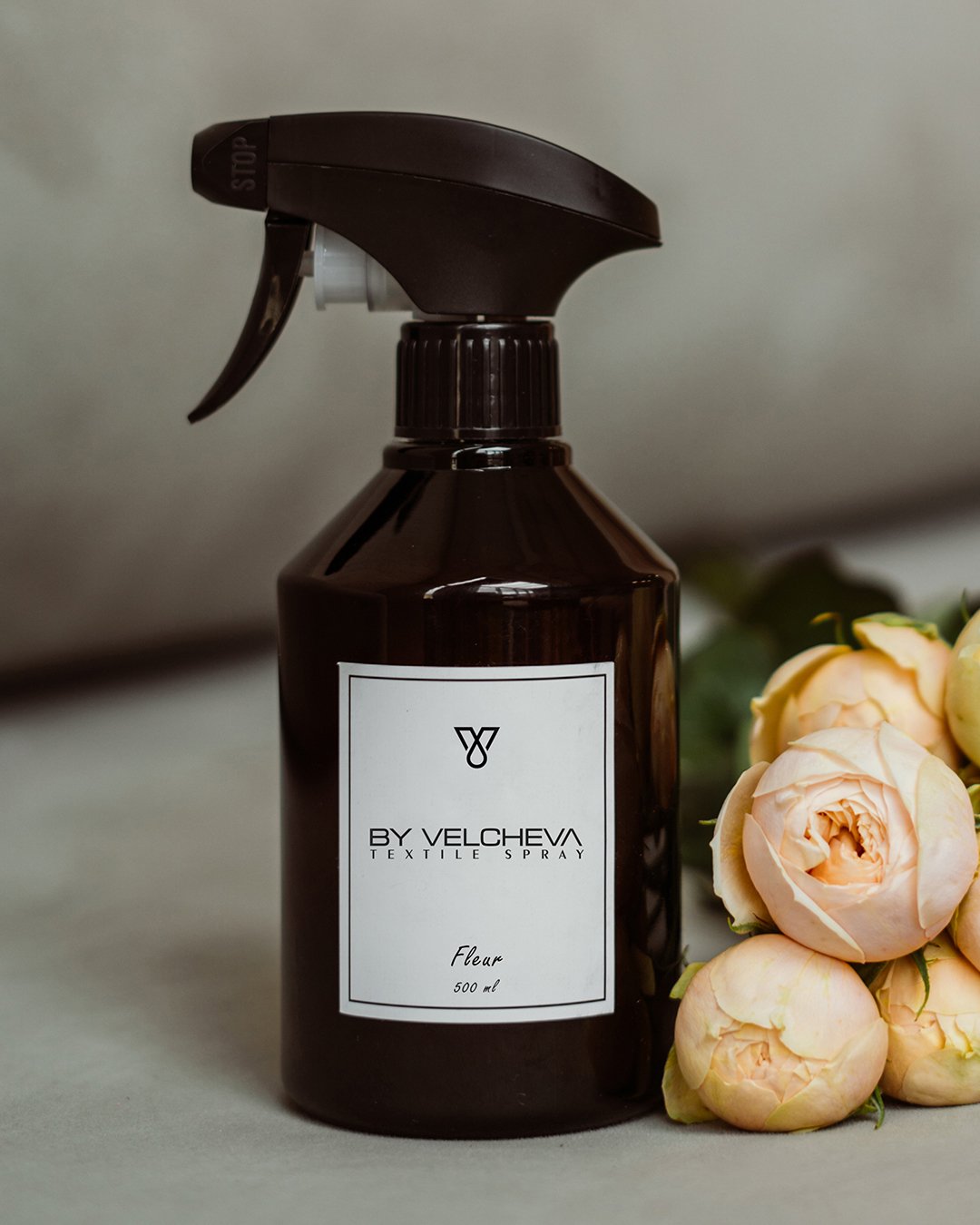 Αρωματικό σπρέι υφασμάτων Fleur BY VELCHEVA 500ml σε καφέ μπουκάλι με αντλία ψεκασμού, δίπλα σε απαλά ροζ τριαντάφυλλα